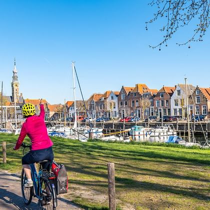 Two cyclists in pink jackets riding along a green waterfront path in Zeeland. Historic Dutch houses and a marina with sailboats visible across the water.