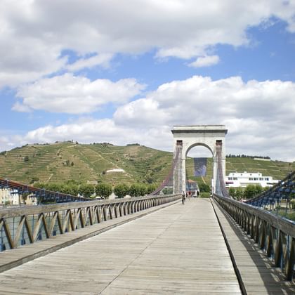 Wooden suspension bridge with metal railings crossing the Rhône river, with terraced vineyards on hillside in background under cloudy sky.