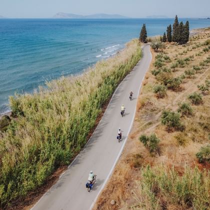 Coastal cycle path in Peloponnese, Greece Aerial view of cyclists on a paved coastal path in Peloponnese, Greece, running between turquoise sea and Mediterranean vegetation.