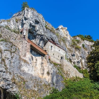 Stone building with red roof built into the white limestone cliff face of Isteiner Klotz, surrounded by green vegetation under blue sky.