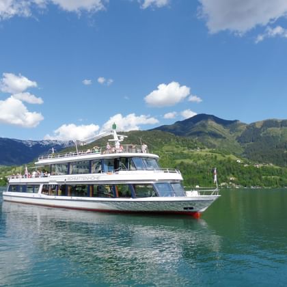 White passenger boat on turquoise lake waters at Zell am See with green mountains and blue sky with white clouds in background.
