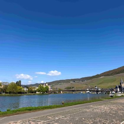 Panoramablick auf Bernkastel-Kues an der Mosel mit Kirchturm, Brücke, Booten und weinbergbedeckten Hügeln unter blauem Himmel.