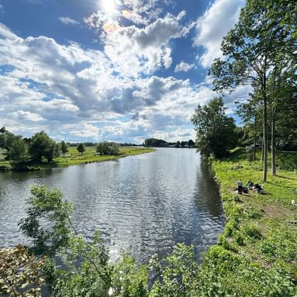 Dove Elbe river flowing through green landscape near Geesthacht. People rest on grassy riverbank under trees, with blue sky and white clouds above.