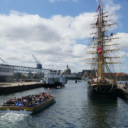 Tour boat passing a tall ship docked in Copenhagen's Christianshavn harbor. Red brick warehouses and a domed church visible in the background.