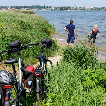 Zwei Radfahrer stehen am Wasserrand eines Ostseestrandes mit Tourenrädern und Packtaschen am grasbewachsenen Ufer. Stadt sichtbar.