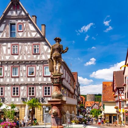 Historischer Brunnen auf dem Besigheimer Marktplatz, umgeben von traditionellen Fachwerkhäusern mit roten Fensterläden unter blauem Himmel.