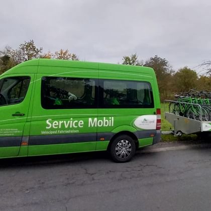 Green van labeled 'Service Mobil' pulling a trailer loaded with multiple bicycles, parked on a road with trees in the background.