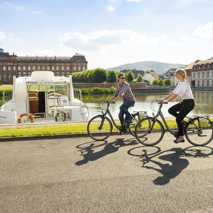 Two cyclists riding along the river port in Saverne, Alsace. A boat is moored on the canal, with historic buildings in the background.