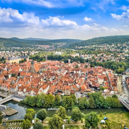 Aerial view of Hann. Münden showing historic town center with red-tiled roofs, river, bridge, and surrounding forested hills under blue sky.