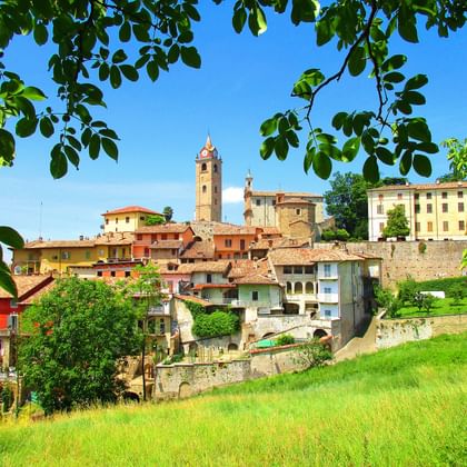 Malerischer Blick auf eine historische Stadt im Piemont mit bunten Gebäuden, einem markanten Glockenturm und grüner Wiese im Vordergrund.