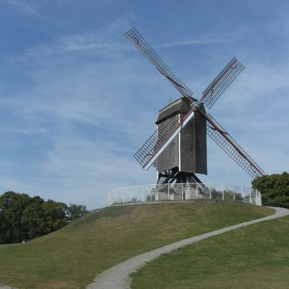 Sint-Janshuismolen Windmühle auf Grashügel Traditionelle Holzwindmühle Sint-Janshuismolen mit vier Flügeln auf einem Grashügel, umgeben von Bäumen unter blauem Himmel mit weißen Wolken.