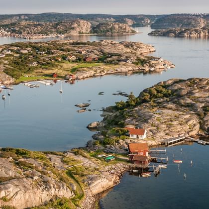 Luftaufnahme eines schwedischen Westküsten-Fischerdorfs mit roten Holzhäusern auf felsigen Inseln, ruhigem Wasser und Booten in der Bucht.