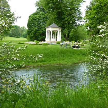 Klassischer Pavillon im Schlosspark Tietfurt Weißer klassischer Pavillon mit Säulen im Schlosspark Tietfurt, umgeben von grünen Rasenflächen und alten Bäumen, mit Bach im Vordergrund.