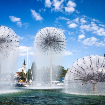 Three large fan-shaped water fountains in Romanshorn spraying water into Lake Constance, with a church tower and marina visible in the background.