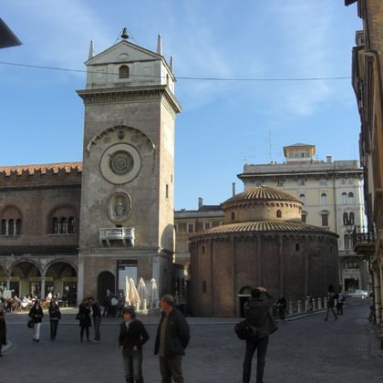 Historic clock tower with large circular clock face dominates Piazza dell Erbe in Mantua, surrounded by medieval buildings and people walking.