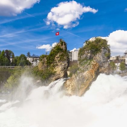 Rhine Falls at Neuhausen with powerful white water cascading over rocks. Swiss flag on rocky outcrop, buildings and blue sky visible.