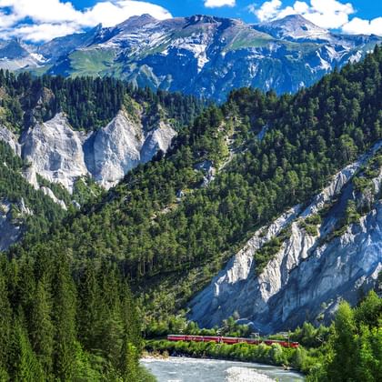 Panoramic view of the Swiss Rhine Gorge showing white limestone cliffs, dense green forests, a winding river, and snow-capped Alps.