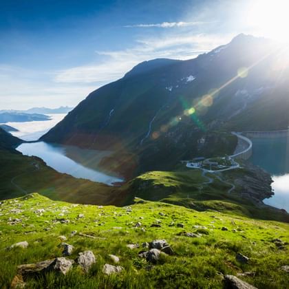 Alpine reservoir in Kaprun surrounded by green slopes and steep mountains. A winding road leads to buildings at the water's edge under bright sunlight.