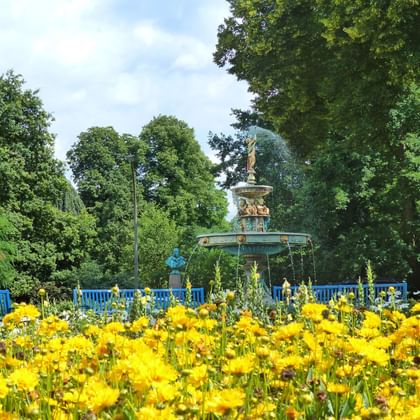 Ornate fountain with blue-green patina in a park surrounded by yellow flowers and blue railings, with tall trees in the background.