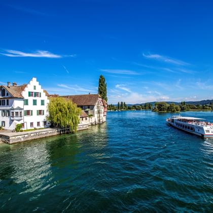 Tour boat on the Rhine River passing historic buildings with painted facades and a church spire in Stein am Rhein under blue sky.
