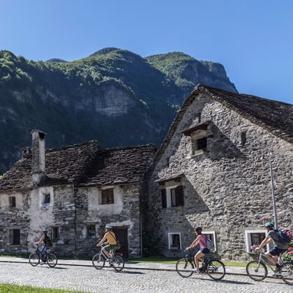 Group of cyclists riding through traditional stone village in Vallemaggia, Tessin, with historic buildings and green mountains in background.