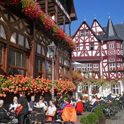 Straßencafé mit Gästen an Tischen vor bunten Fachwerkhäusern in Bacharach, geschmückt mit roten Blumen.