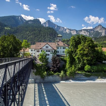 Metal bridge crossing muddy river with Swiss Alpine village and dramatic mountain peaks in background under blue sky with white clouds.