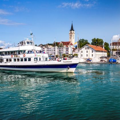 White passenger ferry in Romanshorn port with church tower and historic buildings in background under blue sky with white clouds.