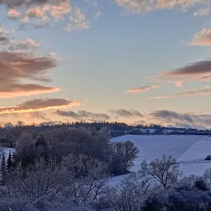 Panoramic winter landscape with snow-covered fields and bare trees under a colorful sunset sky with orange and blue clouds.