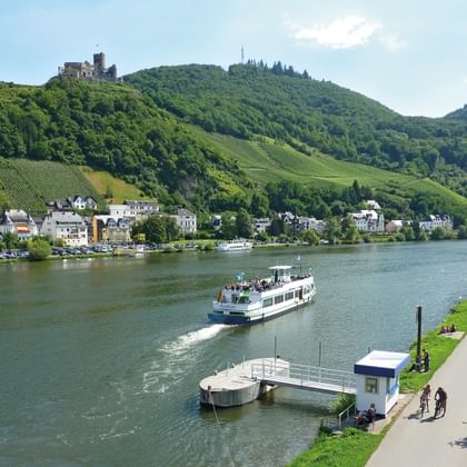 White passenger boat on Moselle River near Bernkastel-Kues with vineyard-covered hills, historic castle ruins, and riverside promenade.