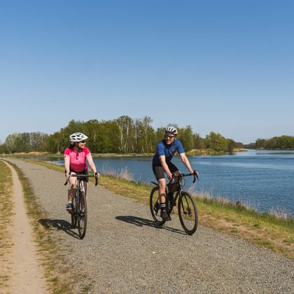 Two cyclists with helmets riding on a paved path beside a large lake. Woman in pink shirt and man in blue shirt cycling under clear sky.