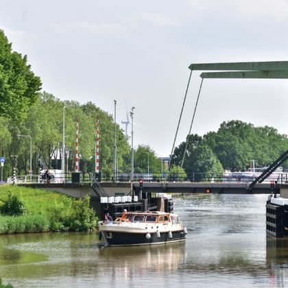 Ein Boot fährt durch einen Kanal unter einer geöffneten grünen Zugbrücke in Vianen, Holland. Grüne Bäume säumen beide Seiten der Wasserstraße.