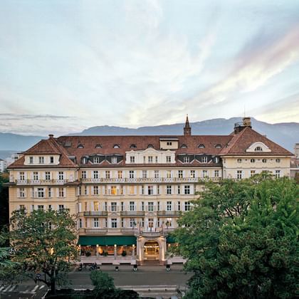 Historisches Parkhotel Laurin Gebäude mit gelber Fassade und rotem Ziegeldach, umgeben von grünen Bäumen mit Bergen im Hintergrund bei Dämmerung.
