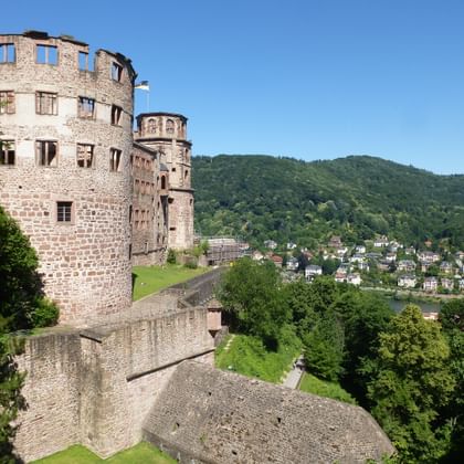 Historic Heidelberg Castle ruins with red sandstone towers and walls overlooking the Neckar River valley with green hills and town below.