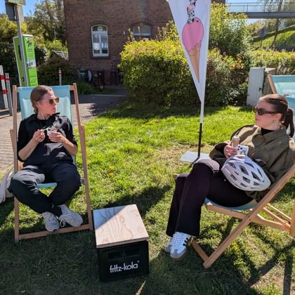 Zwei Frauen entspannen in Liegestühlen auf Gras und essen Eis. Fahrradhelme liegen in der Nähe. Rosa Eisflagge und Backsteingebäude im Hintergrund.