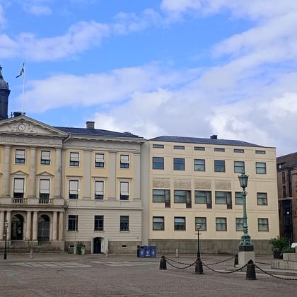 Gustaf Adolfs Torg in Göteborg mit historischem gelben Gebäude, Glockenturm und Bronzestatue auf dem Platz unter teilweise bewölktem Himmel.