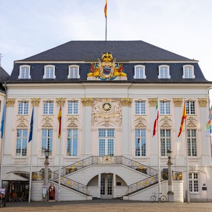 Historic Bonn City Hall with white baroque facade, ornate golden decorations, curved double staircase, and colorful flags on the market square.