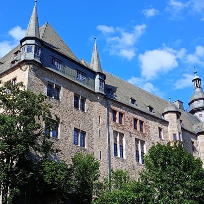 Historic Marburg Castle south wing with stone walls, multiple towers, and conical roofs surrounded by green trees under a blue cloudy sky.