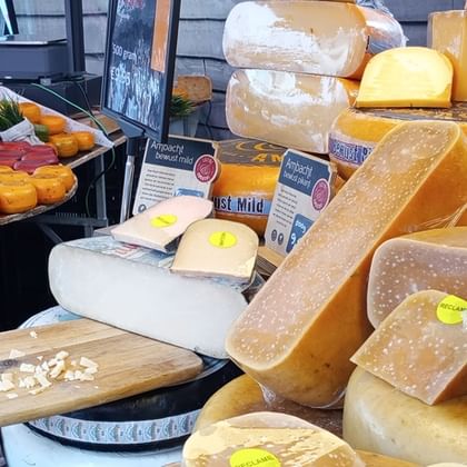 Market stall displaying various Dutch cheese wheels and blocks in Middelburg, Zeeland. Yellow cheese rounds are arranged on trays with larger wheels stacked behind.