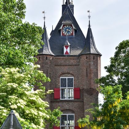 Medieval brick tower with conical slate roofs, clock face, and red shutters surrounded by green trees in a Dutch town setting.
