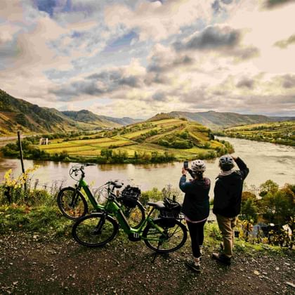 Two cyclists with bikes viewing the Mosel river loop from a hilltop. The river curves around a green peninsula with vineyards and hills in the background.