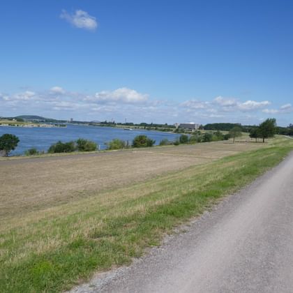Paved bike path running alongside the Ruhr river between Duisburg and Essen, with green fields, trees, and blue sky with white clouds.