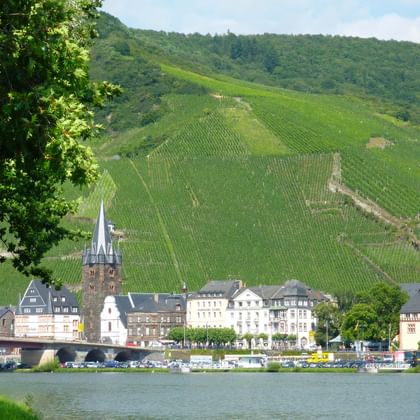 Historic town of Bernkastel-Kues along the Moselle River with traditional buildings, church spires, and steep vineyard slopes rising behind.