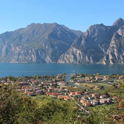 Erhöhter Blick über den Gardasee mit einer Seestadt mit roten Dächern zwischen dramatischen Kalksteinfelsen und blauem Wasser.