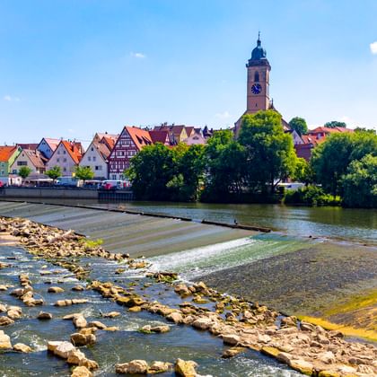 Blick auf Nürtingen mit Kirchturm und historischen Fachwerkhäusern am Neckar. Steiniges Flussbett im Vordergrund, blauer Himmel.