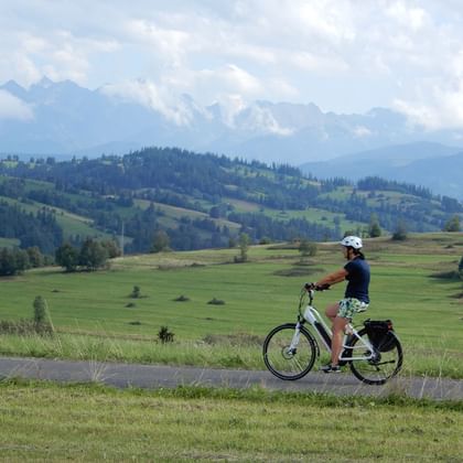 Female cyclist on paved path through green meadows with forested hills and High Tatra mountain range in background under cloudy sky.