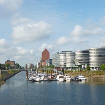 Duisburg harbour showing a canal with moored boats, modern curved buildings on the right, industrial structures on the left, under a cloudy sky.