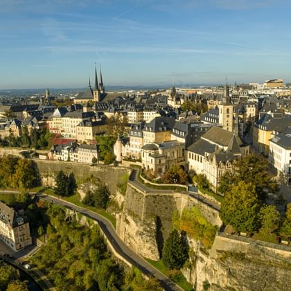 Luftaufnahme von Luxemburgs Chemin de la Corniche mit historischen Festungsmauern, Altstadtgebäuden und Kirchtürmen unter blauem Himmel.
