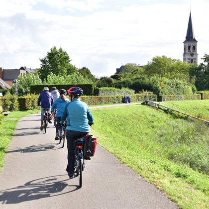 Group of cyclists with panniers riding on paved path through green countryside near Sint-Amands, with church tower visible in background.