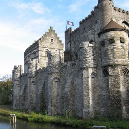 Gravensteen Burg in Gent mit Flaggen Mittelalterliche Burg Gravensteen in Gent mit Steinmauern, runden Türmen und Flaggen auf den Zinnen, am Wassergraben unter blauem Himmel.